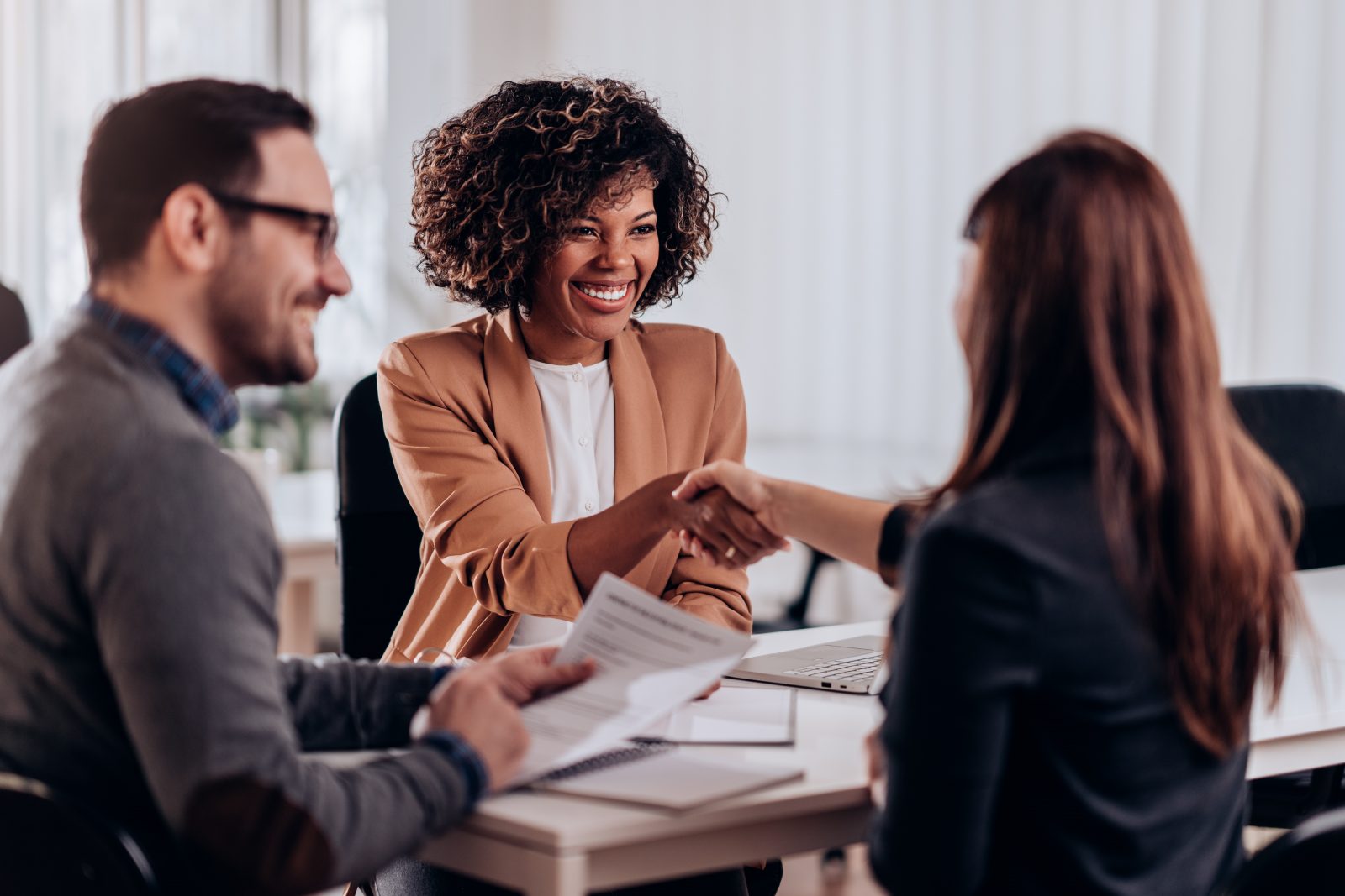 A lady is welcomed to a boardroom by another lady and a man. All are dressed in professional clothing.